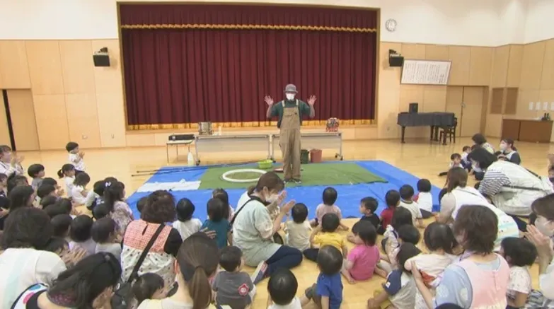 保育園の遊戯室で、ステージ前に座った園児と先生に手を振るシャボン玉おじさん
