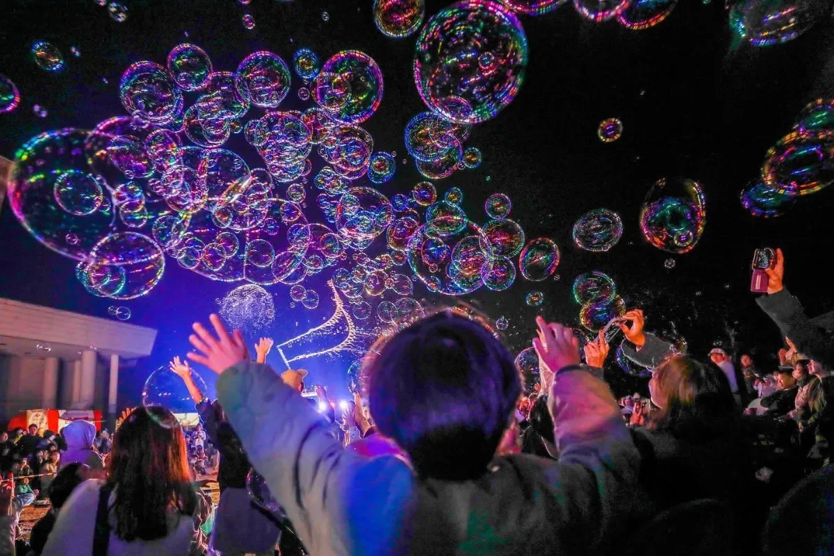 夜空いっぱいに舞う光のシャボン玉と、手をのばして見上げる観客